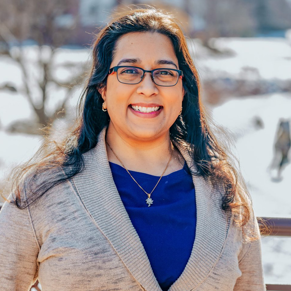 The image shows a woman smiling at the camera, wearing glasses and a blue top, standing outdoors with trees behind her.