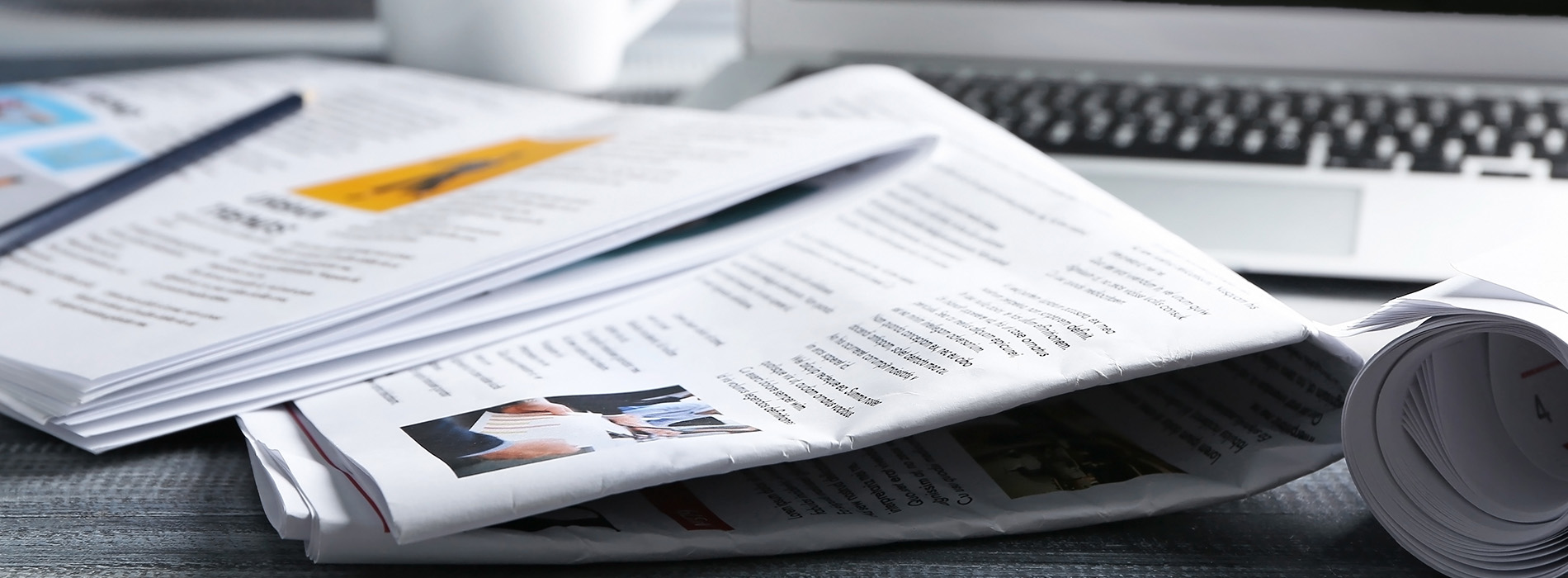 The image shows a collection of newspapers spread out on a surface with a focus on their headlines and front pages, some of which are partially obscured by other papers.
