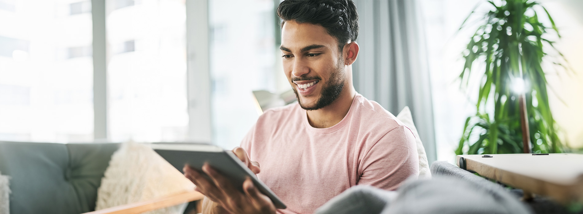 A young man seated on a couch with his hands holding a tablet device, smiling at the camera.
