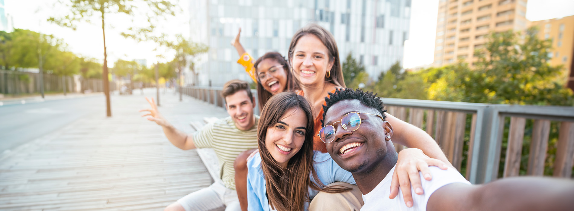 The image shows a group of four people posing for a photo on a wooden deck with a cityscape in the background, one person is taking the picture, they are smiling and appear to be enjoying themselves.