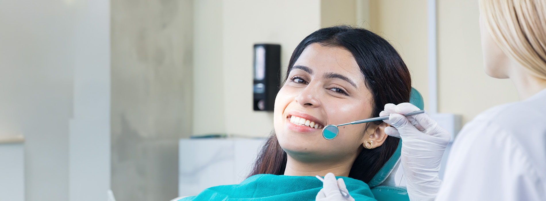 A woman sitting in a dental chair with a smile on her face, receiving dental care from a professional who appears to be taking an x-ray of her teeth.