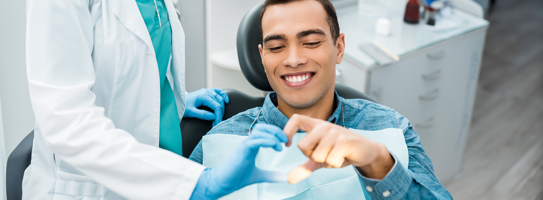 The image shows a dental office setting with a smiling man seated in a dentist s chair, receiving dental care from a professional wearing blue gloves.