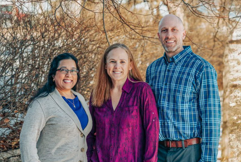 The image shows three individuals posing together outdoors during daylight  they appear to be at an event, possibly a formal occasion given their attire, with one person wearing a tie.