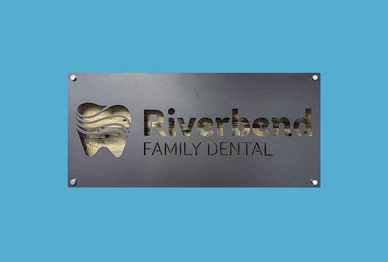 The image features a sign with text that reads  RIVERBEND FAMILY DENTAL  on a blue background, set against a backdrop of a building facade.