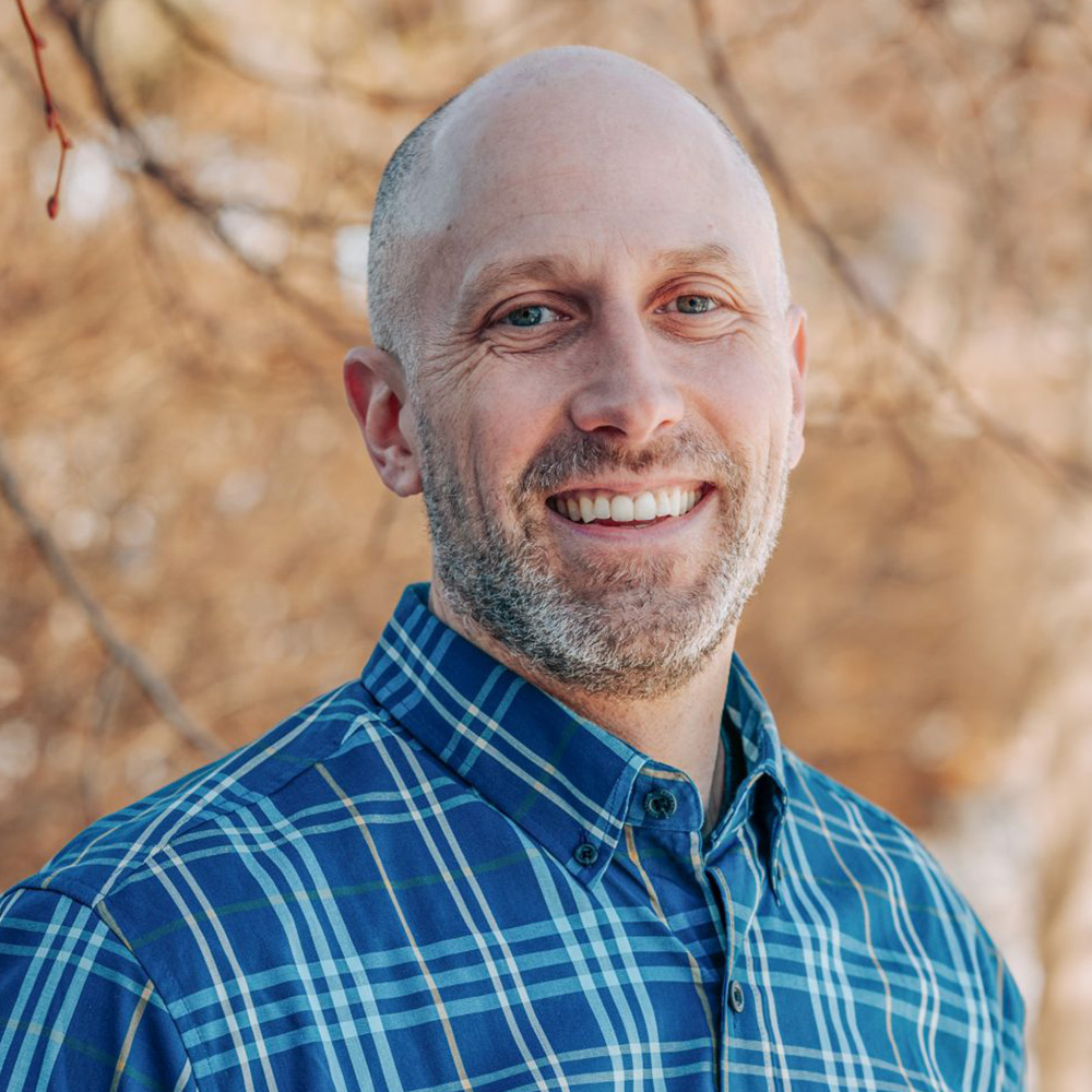 The image shows a man with a beard smiling at the camera, wearing a blue plaid shirt and standing outdoors against a natural background during daylight.
