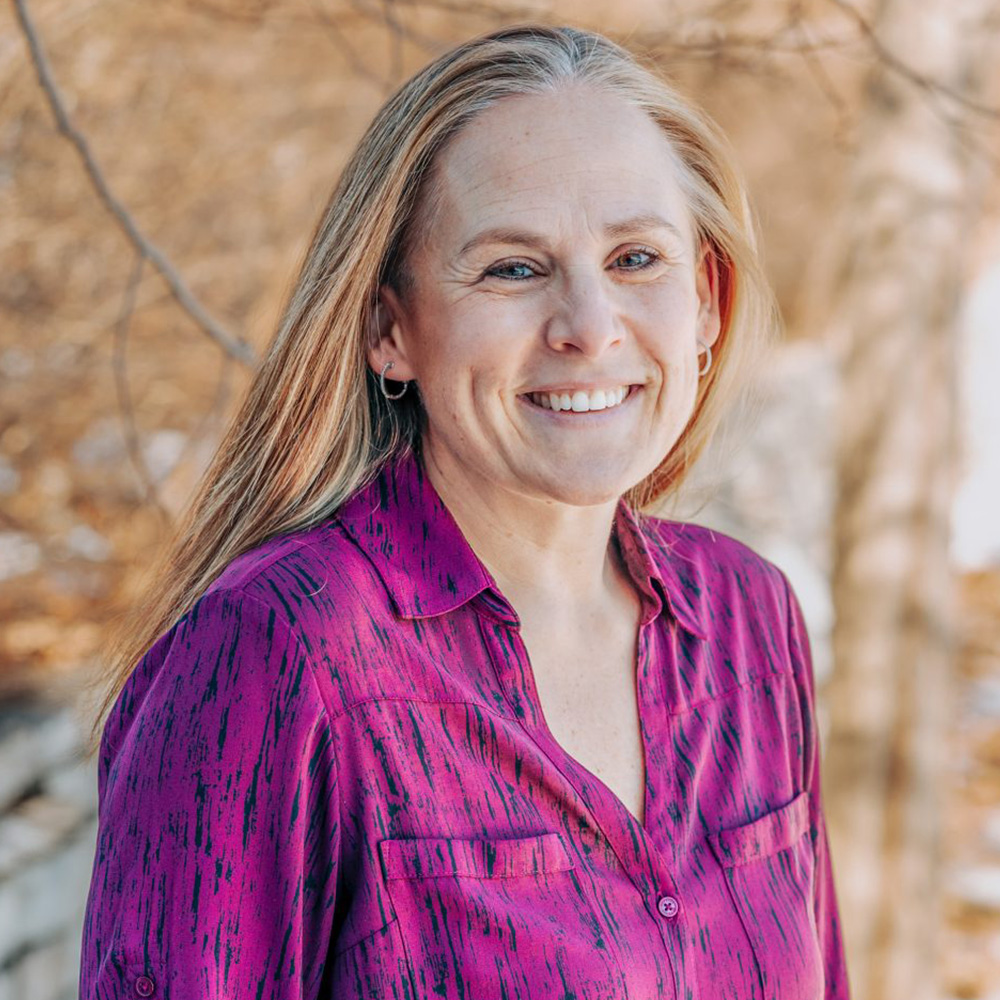 A woman wearing a purple top stands outdoors with trees in the background.