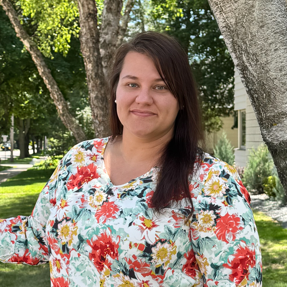 A woman standing outdoors with a floral blouse, smiling at the camera.