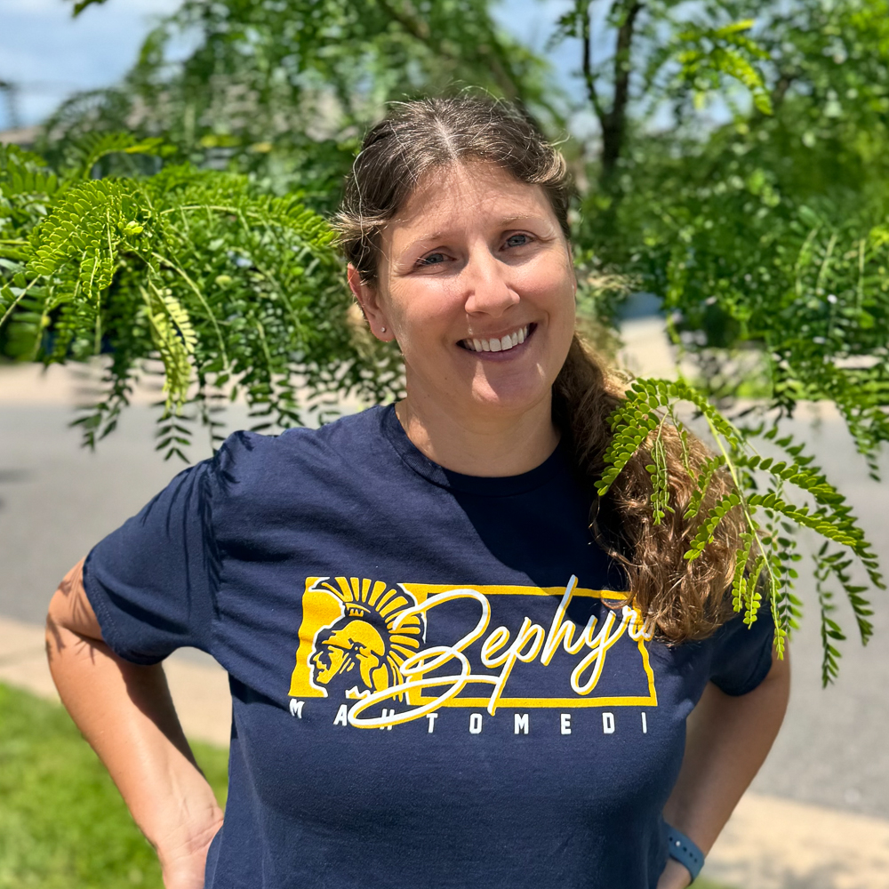 The image features a person standing outdoors with a tree behind them, wearing a dark blue t-shirt with text and logos, posing for the camera with a smile.
