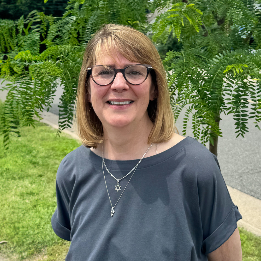 The image shows a woman standing outdoors with her hands by her sides, wearing glasses, a dark top, and a necklace. She has short hair, is smiling slightly, and appears to be posing for the photo. Behind her is a tree with green leaves and a clear sky in the background.