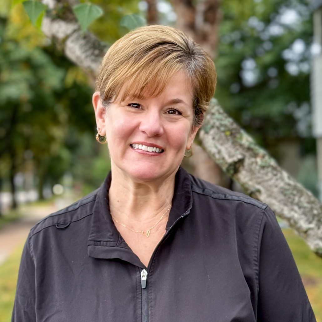 The image shows a woman posing outdoors with a smile, wearing a black jacket and standing next to a tree.