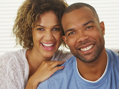 The image shows a man and woman smiling at the camera  the man has a beard and is wearing a blue shirt, while the woman has short hair and is dressed in white. They appear to be in a joyful mood.