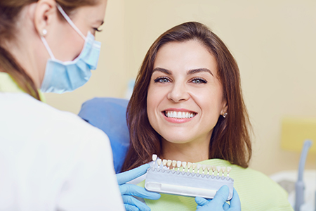 The image shows two women  one sitting with a smile, receiving dental care from another woman who is standing over her, holding a dental device.