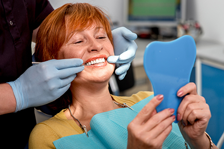 The image shows a woman with red hair sitting in a dental chair, smiling at a blue tooth model held up by a dental hygienist wearing gloves.
