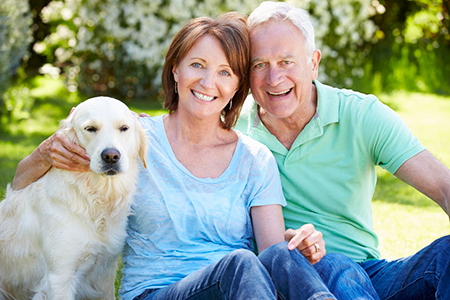 An elderly couple sitting outdoors with a dog on their laps, posing for a photo.