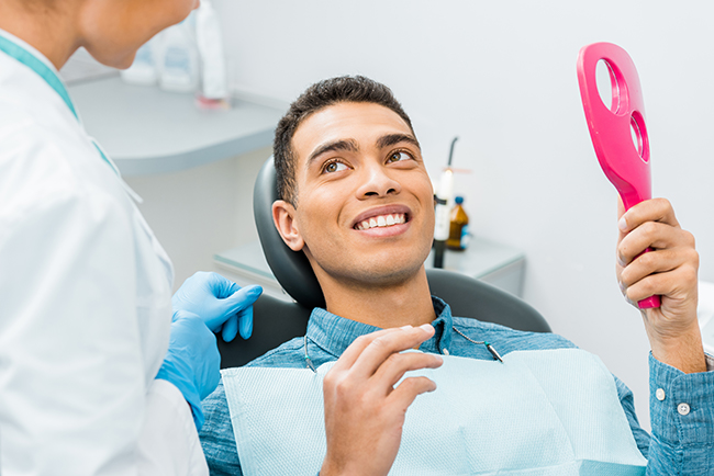 The image shows a man sitting in a dental chair with a smile, holding a red object up to his mouth, while a dental professional attends to him in a dental office setting.
