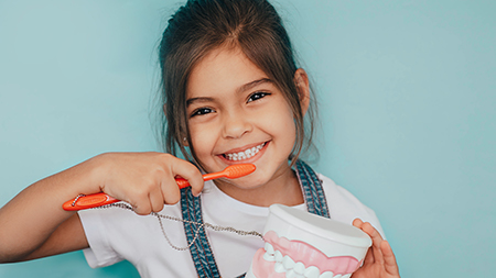 The image shows a young girl brushing her teeth with toothpaste in her hand while smiling at the camera.