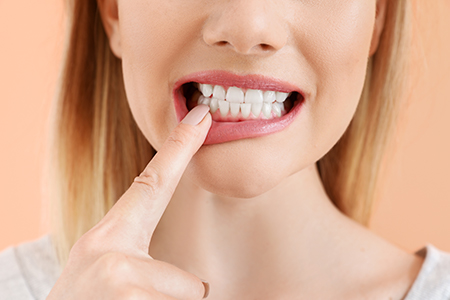 The image shows a woman with her finger in her mouth, pressing on her teeth, against a plain background.