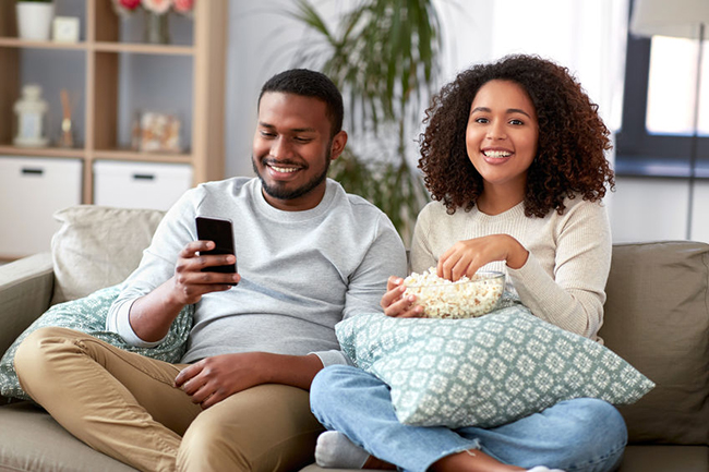 A man and woman are sitting on a couch, enjoying popcorn while watching television together.